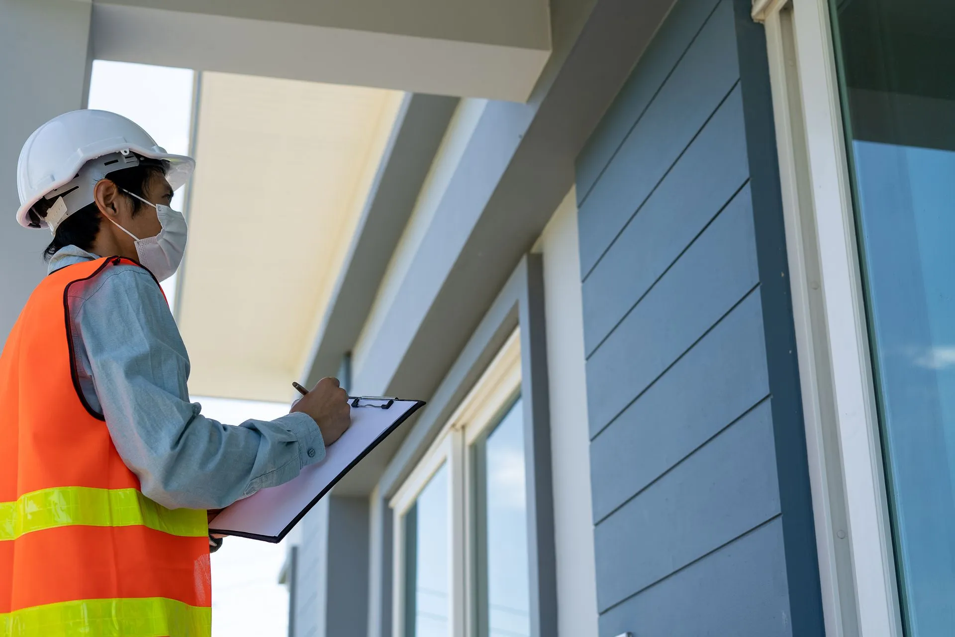 A construction worker wearing a mask and hard hat is looking at a building while holding a clipboard.